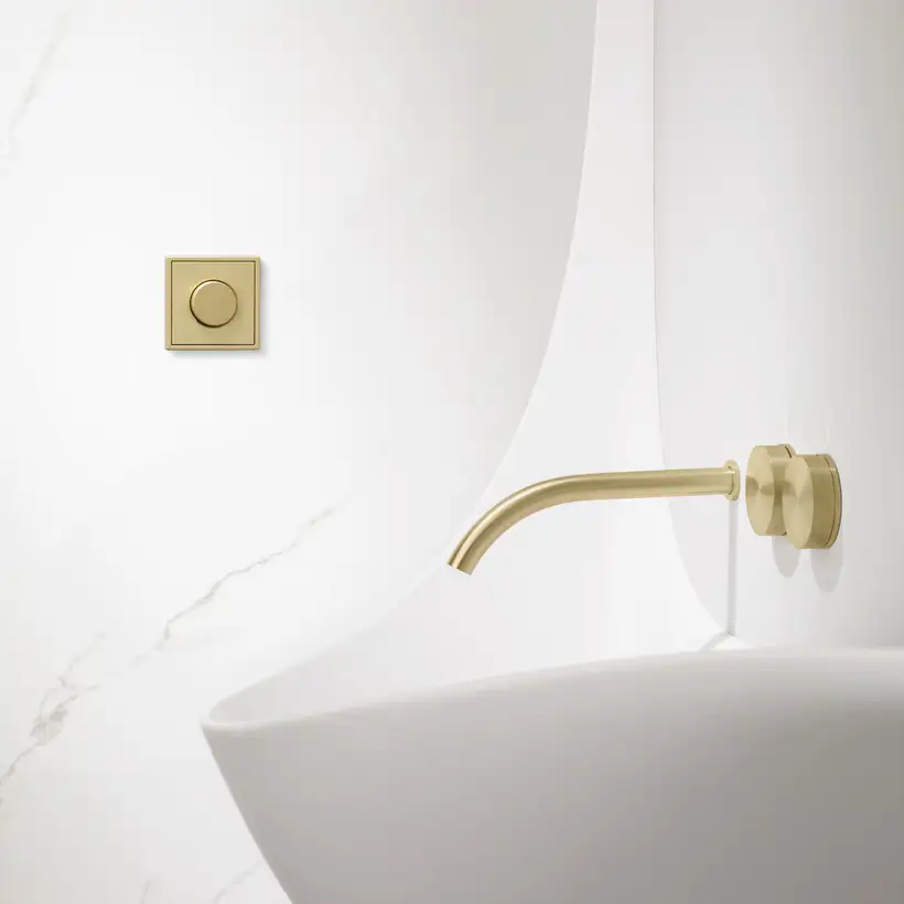 Drehdimmer im Programm LS 990 in Messing Classic, frontal aufgenommen, Raumausschnitt eines Badezimmers mit weißen Marmorwänden, einem geschwungenem weißen Waschbecken und einer schlanken Amatur in gebürstetem Messing.