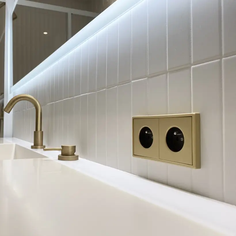 SCHUKO® Steckdosen im Programm LS 990 in Messing Classic, in leichter Perspektive fotografiert, auf weißen Kacheln über einem ebenfalls weißem Waschbecken.