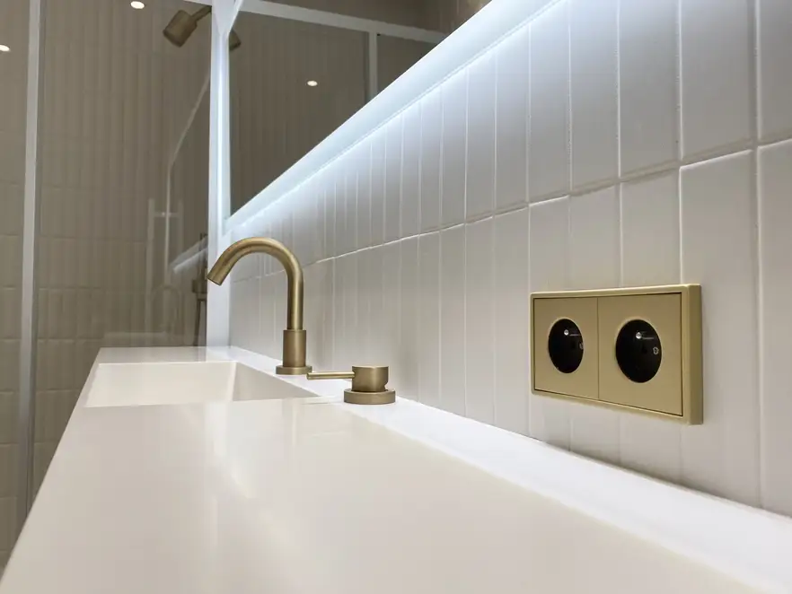 SCHUKO® Steckdosen im Programm LS 990 in Messing Classic, in leichter Perspektive fotografiert, auf weißen Kacheln über einem ebenfalls weißem Waschbecken.