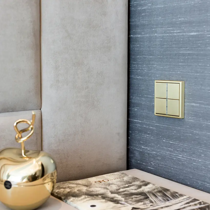 Detailansicht KNX F 40 Taster in Messing Classic, fotografiert in leichter Perspektive, auf einer grauen Wand mit Strucktur. Im Vordergrund wurde ein Katalog und ein goldfarbener Zierapfel dekoriert. 